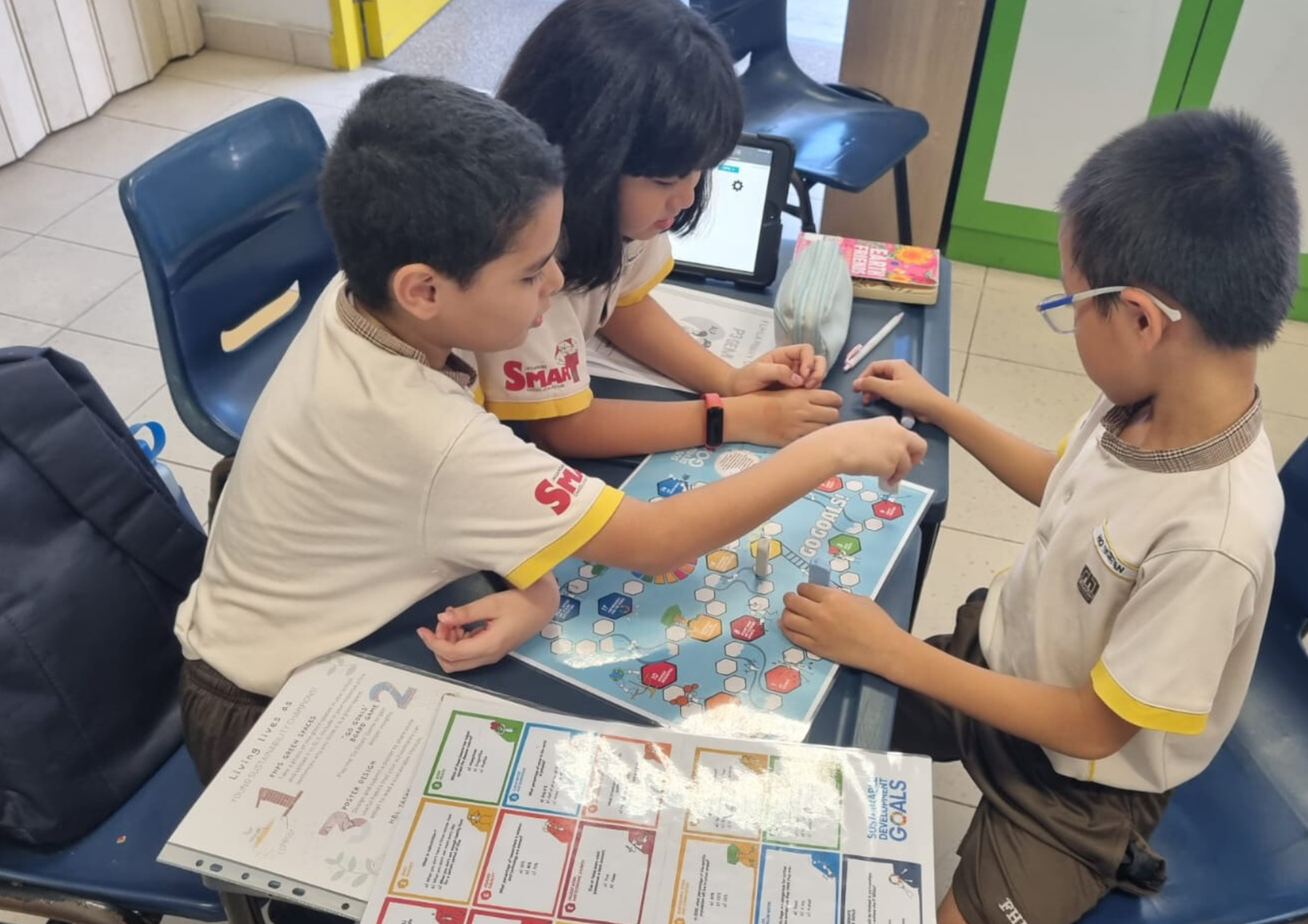 Three children in school uniforms sit at a desk playing a colorful board game called Go Goals.